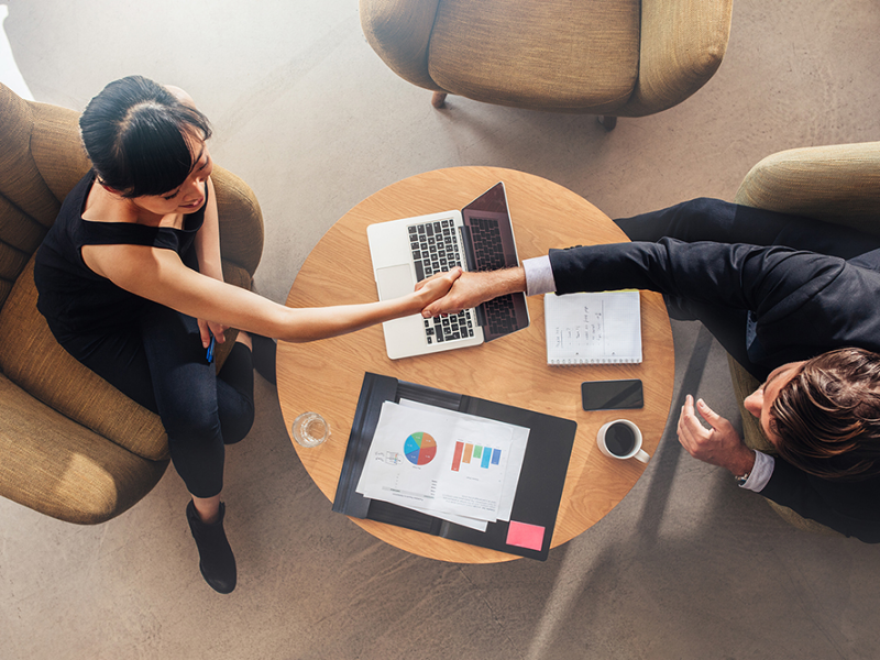 an Asian businesswoman shaking hands with a white businessman in an office setting with laptops and coffee